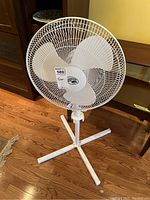 Front view of the white Hampton Bay standing fan showing the blades, protective grille, and cross-shaped base on a hardwood floor.