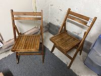 Two matching wooden folding chairs with slatted seats and backs positioned upright against a wall on carpeted floor.