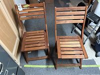 Pair of two folding wood chairs, showing their front and back views side by side on a carpeted floor.