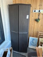 Front view of tall resin garden shed with woven lattice style doors, sitting outdoors with wooden wall in the background and boxes nearby.