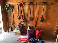 Photo showing multiple gardening tools hung on wood wall including rakes, shovels, and a Toro electric edge trimmer with gardening hose coiled on wall. Several bags of soil amendments and planters on floor.