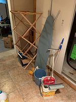 Lot overview showing wooden drying rack, ironing board, mop bucket and mop, red bucket, and white containers.