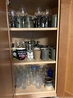Shelf with various stemmed wine glasses, green and beige ceramic mugs, a large black mug with rescue mom text, and small glass jars.