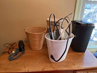 Photo of black plastic waste bin, white metal bucket containing power bars and cords, two wicker baskets on a wooden table near window.