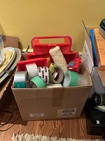 Box on floor containing red plastic paint tray, various rolls of painter's tape, a white paint roller cover, and Scotch tape.