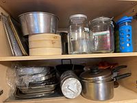 Photo showing shelf with stacked metal pans, bamboo steam baskets, clear glass jars and containers with lids, metal grater, and a reusable water bottle in blue silicone sleeve.