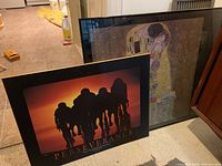 Photo showing both framed prints: a large Klimt 'The Kiss' print behind a smaller Perseverance cycling print with cyclists silhouetted against sunset.