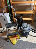 Folding step ladder and gray shop vacuum with hose and filters on concrete basement floor near wooden shelves