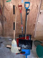 Full lot showing three snow shovels standing against a wooden wall, two brooms, and dustpan with hand brush lying on the floor