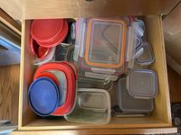 Photo showing a drawer filled with assorted plastic food storage containers with different colored lids, mostly rectangular and square in shape.