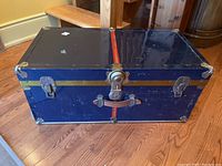 Front view of dark blue heavy metal trunk with metal trim, two latch closures, central lock, and worn leather handle.