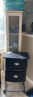 Photo showing both black wheeled bath storage unit with two cloth drawers and tall cream-colored bath cabinet with glass door.