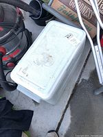 White hard plastic Coleman cooler with scuff marks sitting on garage floor among other items.