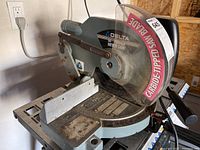 Side view of the used Delta 10 inch power miter saw with clear protective blade guard and angle scale on base. Shows rust on arms and base.