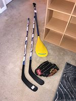 Two Colorado Avalanche hockey sticks, Airhead aluminum paddle, and Rawlings mitt shown on carpeted floor near wooden shelving.