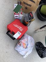 Lot items arranged on concrete including Milwaukee red case, bucket with nails, digital deadbolt box, and a plastic bag with hand warmers.