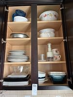 Four shelves of kitchenware in dark wood cabinet including bowls, plates, baking dishes, and kitchen tools