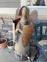 Full side view of metal angel showing smooth head, clasped hands, textured wings, and scroll patterned midsection with star designs on base.