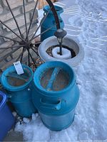 Three teal painted metal milk cans with visible rust and chips in paint next to a concrete planter with a black solar light inside, outdoor setting with snow.