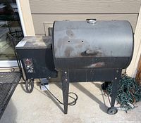 Exterior of black Traeger pellet grill smoker on four legs (two wheels, two fixed), side pellet hopper and control panel, surface rust on lid.
