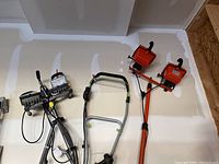 Two tripod mounted sets of work lights and three halogen lights mounted on an orange metal board against a garage wall, cords loosely coiled and hanging.