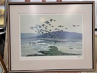 Framed lithograph featuring geese flying over wetlands with a mountain background.