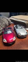 Two diecast metal convertible cars placed on a wooden surface with a cat nearby.