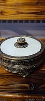 Closed view of the round bronze trinket box featuring the white glass lid inset with ornate finial knob on top and decorative embossed sides.