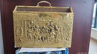 Front and side view of the vintage brass-engraved wooden magazine holder showing detailed figures and foliage engraving on brass panels, along with the brass handle on top.