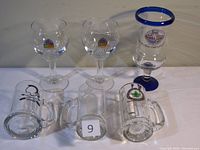 Six beer glasses arranged in two rows on white surface. Two clear goblet style Leffe glasses, one clear bubbled stemmed Bubba Gump glass with blue rim, three clear mugs including one with military insignia, one with festival logo, one plain.