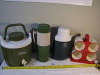 Photo showing all the camping beverage containers from Coleman, Thermos, and Charwood including the water jug, two vacuum bottles, and cup holder with mugs