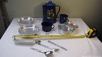 Photo showing all items spread on a table with tape measure for scale: two metal plates, blue enamel jug, two cups, metal pot with lid, metal cutlery set including spoon, fork, knife, and a clear glass jar with metal lid.