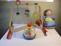 Wooden baby gym with hanging toys, Playscool spinning top, and wool doll shown together on white surface.