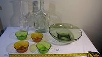 Lot of glass items on white cloth including large green bowl, four small green and amber bowls, two clear glass pitchers with one marked 'white', and a decorative clear decanter.