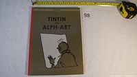 Front cover of the Tintin and Alph-Art book with a grey background, red spine, and a black line drawing of a person holding a paper.