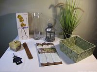 Photo showing the clear glass vase, metal lantern, decorative plastic plant, woven basket, candle, wooden incense holder, and incense packets arranged on a white surface against a wall.