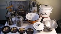 Photo showing assortment of metal and enamel kitchenware items arranged on a table, viewed from front angle.