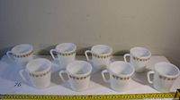 Eight white Pyrex coffee cups with gold floral band, arranged in two rows showing all cups clearly.