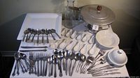 Photo showing the assortment of white porcelain, ceramic, and metal cutlery laid out on a table including plates, spoons, forks, chafing dish, and carafe.