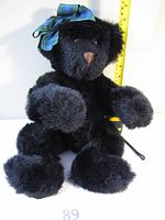 Front view of the black teddy bear sitting upright, showing the plaid bow on its head and plush brown nose.