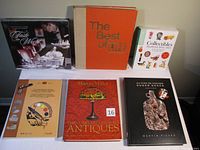 Six books arranged upright and flat on a surface, showing front covers and spines. Titles visible include a culinary book sealed in plastic, an orange and beige large book, an antiques guide with a red cover, and a signed cookbook with a black cover featuring food imagery.