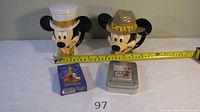 Two Mickey Mouse coin banks, one with a white sailor hat and one with a safari hat, alongside two decks of playing cards on a white surface with a measuring tape indicating approximately 7 inches tall.