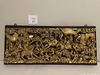 Full front view of the gold painted wood carving depicting warriors on horses in a battle scene with detailed surrounding foliage and fortress in background.