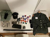 Full overview of the vintage US Army Class A uniform pieces including jacket, pants, caps, military patches, pins, belt, and socks spread out on a table.