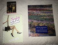 Three books displayed on bed: small hardcover Rembrandt art book, paperback Clark Art Institute list book with color cover, spiral-bound crossword book with polka dot cover and person on swing illustration.