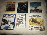 Top view of all six books arranged on a carpeted floor: Truckers Atlas, The Solar System, Volcano, Flight, The Way Things Work CD-ROM, Explore the World of Man-Made Wonders.