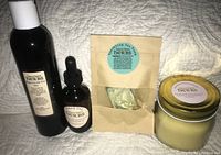 Photo showing all four products: Magnesium Oil Gel in tall dark plastic bottle, Beard Oil in small dark glass dropper bottle, Sparkling Sea Foam soap in tan pouch, Sandalwood Beeswax candle in glass jar with gold lid.