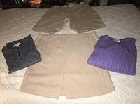 Photo of all four clothing items laid out on a bed: beige shorts on top, beige skirt below, purple sweater to right, gray ribbed shirt to left.