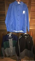Three men's jackets and hoodies displayed hanging on hangers against wood background: NY DEC blue windbreaker top, multicolor 10th Gear jacket below left, and black/gray Starter full zip hoodie below right.