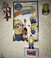 Lot of four character collectible items: Shrek the Third mini Shrek head viewfinder, Blue M&M train figure, Minions Play Pack package, and Music Mate Tim Minion figure shown on quilted background.